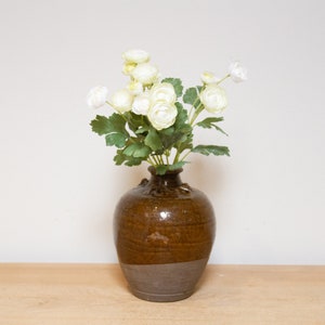 May include: A bouquet of white flowers in a brown ceramic vase. The vase has a textured surface and a wide, round base.
