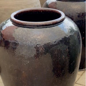 May include: A collection of large, glazed ceramic pots in varying shades of brown. The pots have wide openings and rounded bodies. Some pots have a textured surface, while others are smooth and glossy. The pots are arranged outdoors.