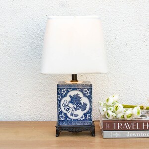 May include: A blue and white porcelain table lamp with a white lampshade. The lamp base features a detailed floral and dragon design. The lamp is on a black stand, next to books and flowers.