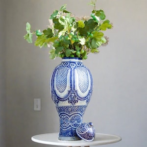 May include: A large blue and white ceramic vase with intricate geometric patterns, filled with green leafy branches and small green berries, sits on a white pedestal table.