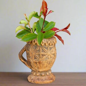 May include: A carved wooden vase with a geometric pattern, featuring a brown and beige color scheme. The vase is filled with green and red leaves.