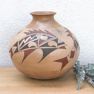 May include: A tan-colored, round ceramic vase with a narrow neck. The vase is decorated with a geometric design in black, red, and white, featuring stylized fish and star-like shapes. The vase sits on a wooden surface.