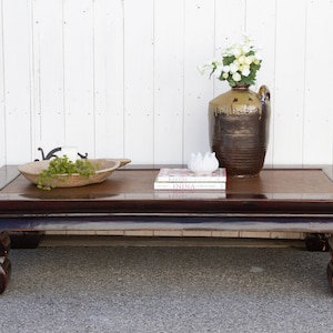 May include: A dark wood coffee table with a rectangular top and four legs. The table has a raised edge around the top and the legs are curved. There is a wooden bowl with greenery, two books, and a brown ceramic vase with white flowers on top of the table.