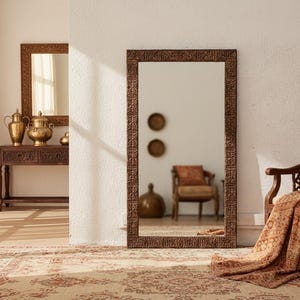 May include: A large, rectangular mirror with a detailed, brown frame stands against a white wall. A wooden chair with an orange patterned throw and a decorative table with gold vases are also visible. The room has a warm, inviting atmosphere.