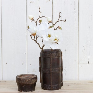 Puede incluir: Un arreglo de flores de magnolia blancas en un jarrón cilíndrico tejido de color marrón oscuro. El jarrón tiene un patrón tejido decorativo y se encuentra sobre una superficie de madera marrón claro.