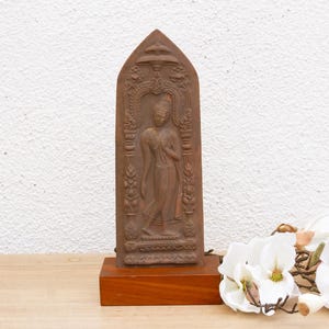May include: A brown wooden sculpture of a standing Buddha figure with intricate carvings. The sculpture is on a wooden base and is displayed against a white wall.