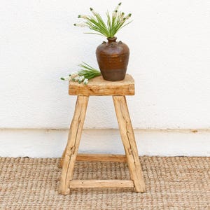 May include: A rustic wooden stool with a natural finish. The stool has a simple design with four legs and a square top. A brown ceramic vase with white flowers sits on top of the stool.