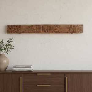 May include: A long, rectangular, wooden wall decor piece with ornate carved floral and scroll designs. The brown piece is mounted on a white wall above a dark brown cabinet with gold handles. A vase with greenery sits on the cabinet.