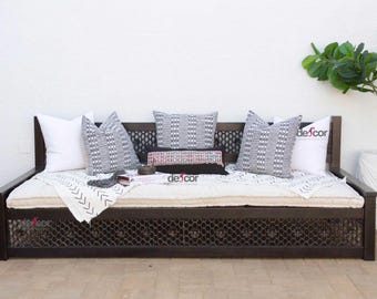 May include: A dark brown wooden daybed with a white mattress and a black and white patterned throw blanket. The daybed has a black metal mesh design on the front and is decorated with white, black, and gray patterned pillows. The daybed is in front of a white wall and a potted plant with green leaves.