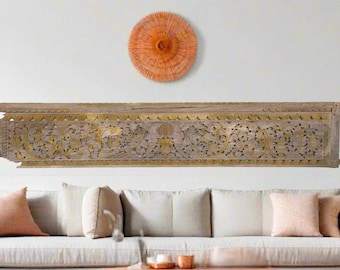 May include: A long, rectangular, carved wooden panel with intricate floral and leaf designs, accented with gold paint. Above, a circular woven wall hanging in shades of orange. The panel is mounted above a white sofa with peach-colored pillows.