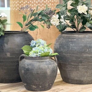 May include: Three dark gray, textured ceramic vases of varying sizes, each holding different greenery and flowers. The largest vase on the right has white flowers, the middle vase has blue and green flowers, and the left vase has green foliage.