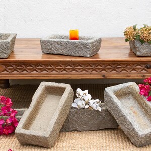 May include: A wooden coffee table with carved details supports several rectangular stone planters. One planter holds a lit orange candle. Other planters are on the floor, some with flowers. The planters are made of a rough, textured stone.