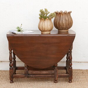 May include: A dark brown wooden drop-leaf table with turned legs and a folded side. On top sit two light brown, petal-shaped decorative vessels, one containing small green sprigs, and a small stack of white papers with a single green stem.