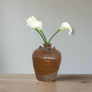 May include: A brown ceramic vase with a textured surface and a wide mouth, holding three white calla lilies with green stems.