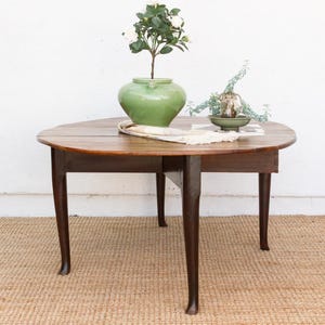 May include: A round, wooden table with dark brown legs and a light brown tabletop. A green vase with white flowers sits on the table, along with a small dish and a decorative cloth. The table is on a woven rug.