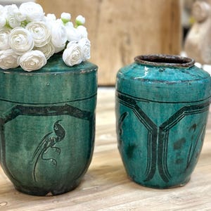 May include: Two turquoise ceramic vases. One vase holds white flowers. The vases have black painted designs, including a bird and geometric patterns. The vases are on a wooden surface.