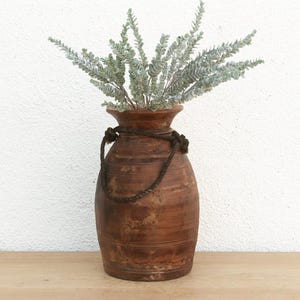 May include: A rustic brown wooden vase with a rope handle, filled with sprigs of silvery-green foliage. The vase has a distressed finish and sits on a wooden surface against a white wall.