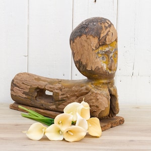May include: A wooden sculpture of a person's head and torso, carved from a tree trunk. The sculpture is painted in shades of brown and yellow. A bouquet of white calla lilies is in the foreground.