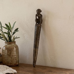 May include: A dark brown metal sculpture featuring a monkey figure perched atop a long, tapered, pointed spike. The object has a weathered, matte finish and stands vertically, showcasing a detailed primate form with arms resting on its knees.