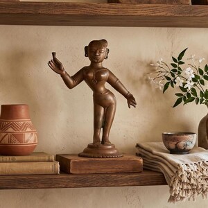 May include: A bronze-toned metal sculpture of a standing figure with one arm raised, positioned on a wooden block. The figure is displayed alongside a terracotta pot with white geometric patterns, stacked books, a small bowl, and a ceramic vase.