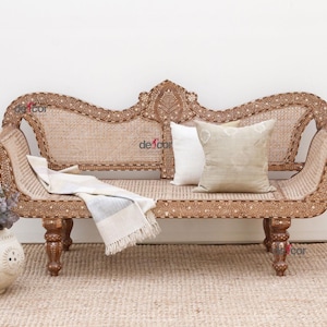 May include: A decorative wooden sofa with intricate carvings and a woven seat. Two pillows and a folded blanket rest on the sofa. A vase with flowers sits to the left of the sofa.