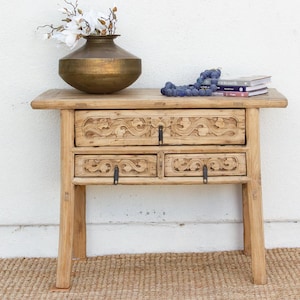 May include: A light-colored wooden side table with two drawers, each adorned with carved floral designs. A brass vase with white flowers sits atop the table, along with a strand of decorative beads and a stack of books. The table is on a woven rug.