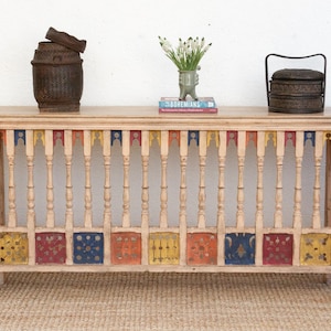 May include: A light wood console table with a painted, decorative, multi-colored, geometric design. The table has a rectangular top and a decorative, open, slatted, railing-style base. The table is decorated with a colorful, geometric pattern. The table is sitting on a light brown rug.