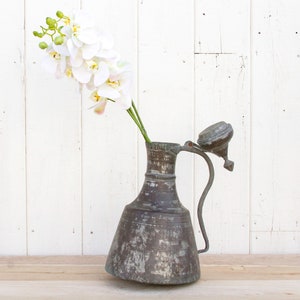 May include: A vintage copper pitcher with a hinged lid and a spout. The pitcher has a worn, aged patina and is filled with a single stem of white orchids.