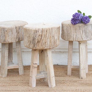 May include: Three rustic wooden stools with a natural, weathered finish. Each stool has a round top and three sturdy legs. One stool is adorned with a cluster of purple flowers. The stools are placed on a woven rug.