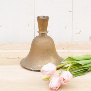 May include: A gold bell-shaped vase with an ornate design sits on a wooden surface with a bouquet of pink tulips.