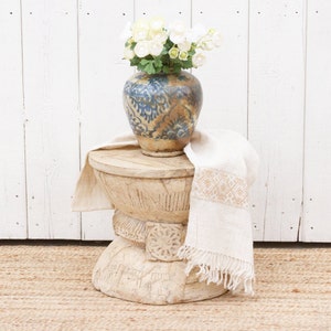 May include: A white wooden stool with intricate carvings sits on a woven rug. A white cloth with fringe hangs over the side of the stool. A blue and white ceramic vase with a floral design holds a bouquet of white flowers.