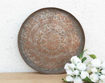 May include: A round, antique copper tray with intricate embossed floral and geometric patterns. The tray has a weathered, aged appearance. A bouquet of white flowers rests on the wooden surface next to the tray.