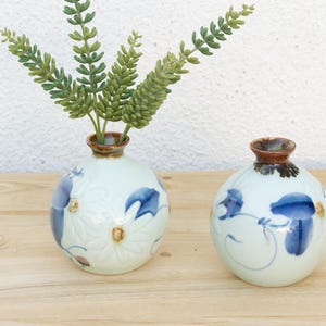May include: Two round ceramic vases with a pale blue-green glaze, featuring hand-painted blue floral patterns and brown-rimmed openings. The left vase contains a green artificial plant with multiple stems and textured leaves.