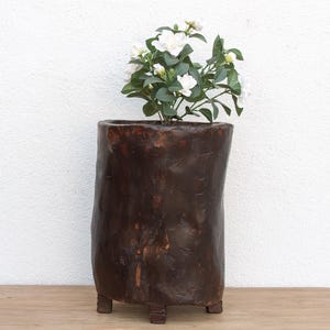 May include: A dark brown, cylindrical wooden planter with a rough, textured surface. The planter sits on three small, square feet. White flowers and green leaves sprout from the top of the planter, creating a natural contrast.