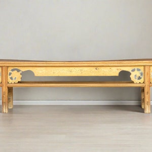 May include: A long, wooden table with a carved design on the front and sides. The table has a light brown finish and is sitting on a light gray floor.