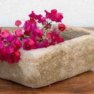 May include: A rectangular stone planter filled with vibrant pink bougainvillea flowers. The planter has a rough, textured surface and sits on a wooden table. The flowers add a pop of color against the neutral tones.