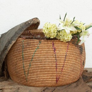 May include: A round, woven basket with a lid, filled with white and yellow flowers. The basket is made of natural fibers and has vertical stripes of green and purple. The lid is made of a dark, textured material.