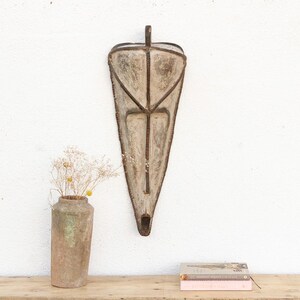 May include: A rustic, weathered wall hanging with a triangular shape and a metal frame. The wall decor is mounted on a white wall, with a vase of dried flowers and a stack of books on a wooden surface below. The piece has a vintage, antique aesthetic.