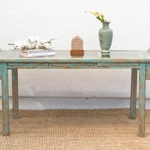 May include: A teal blue wooden console table with a glass top. The table has a distressed finish and intricate carvings on the legs. The table is sitting on a woven rug.
