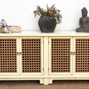 May include: Cream-colored cabinet with four doors featuring a square lattice design. The cabinet is topped with a dark vase holding dried flowers, a small wooden bowl with white flowers, and a seated Buddha statue.