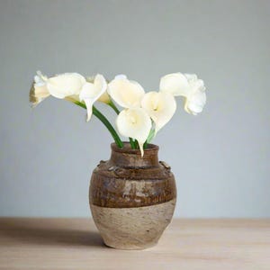 May include: A bouquet of white calla lilies in a brown and beige ceramic vase.