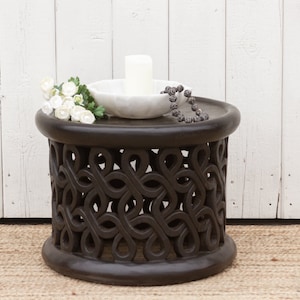 May include: A dark brown wooden stool with an intricate carved design. The stool has a round top and a circular base. A white ceramic bowl with a white candle sits on top of the stool, along with a string of beads.