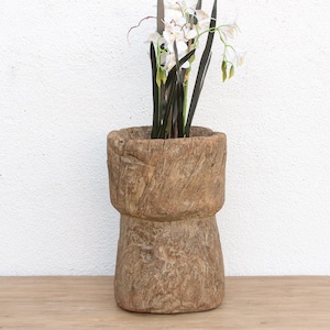 May include: A rustic, wooden vase with a cylindrical shape, featuring a textured surface and natural wood grain. The vase holds artificial white flowers with dark green stems and leaves, creating a natural contrast. The vase sits on a wooden surface.