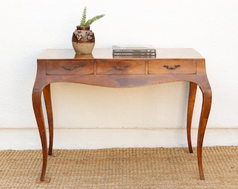 May include: A wooden writing desk with a smooth, polished finish, featuring three horizontal drawers with dark metal handles and curved legs. A small brown ceramic vase with a plant and two stacked books sit on the desk surface.