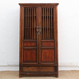 May include: A tall, antique wooden cabinet with two doors. The doors feature vertical wooden slats at the top and solid panels below. The cabinet has a rich, reddish-brown finish and brass hardware. It stands on a simple base.