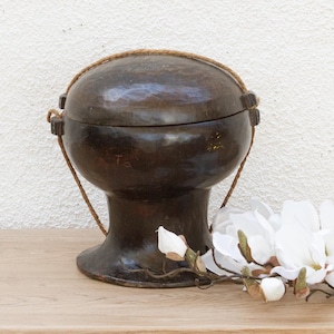 May include: A dark brown, antique-style container with a lid and a woven handle. The container sits on a wooden surface next to a sprig of white flowers. The container has a unique shape and texture.