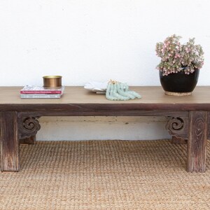 May include: A rectangular, low wooden coffee table with dark brown carved legs and decorative side panels. The table is topped with a light-colored wood surface, and holds a black vase with pink flowers, books, and decorative items.
