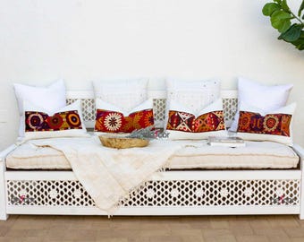 May include: White daybed with a white lattice design on the front. The daybed has a white mattress and is decorated with five pillows. Two of the pillows are white and three are patterned with red, orange, and yellow.