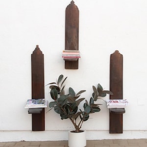May include: Three dark brown wooden wall shelves with a decorative top are mounted on a white wall. The shelves hold books and magazines. A potted plant with dark green leaves sits below the shelves.