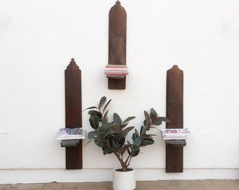 May include: Three dark brown wooden wall shelves with a decorative top are mounted on a white wall. The shelves hold books and magazines. A potted plant with dark green leaves sits below the shelves.